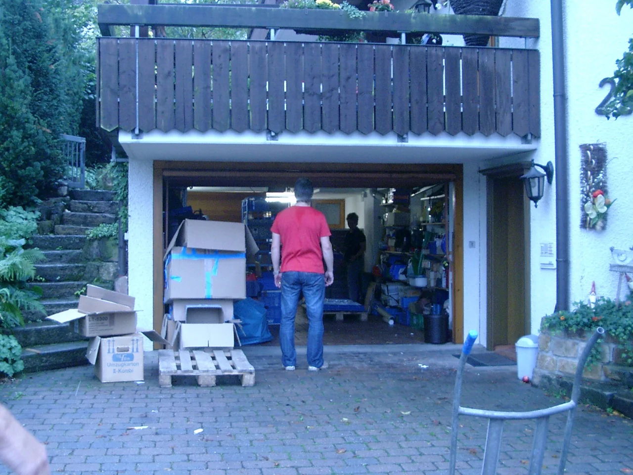 Kromer-GmbH-Oberboihingen-Anfaenge Blick in eine geöffnete Garage mit Kartons, Lagerbehältern und Personen bei der Arbeit – improvisierter Werkstattbereich